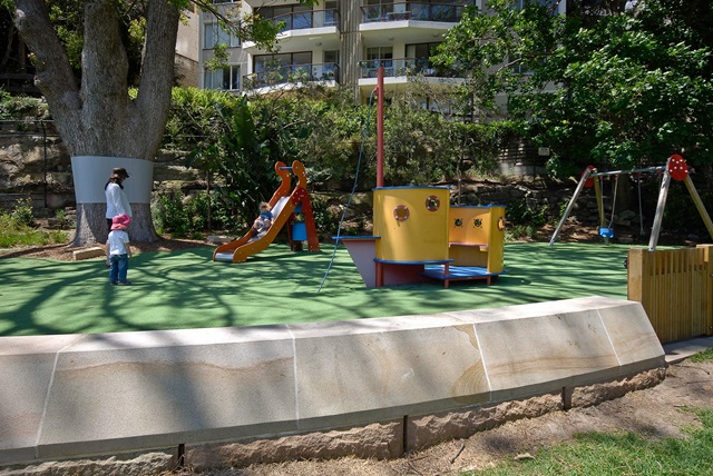 semi enclosed play equipment that resembles a boat. Trees, building and a family watching a kid glide down the slide in the background.